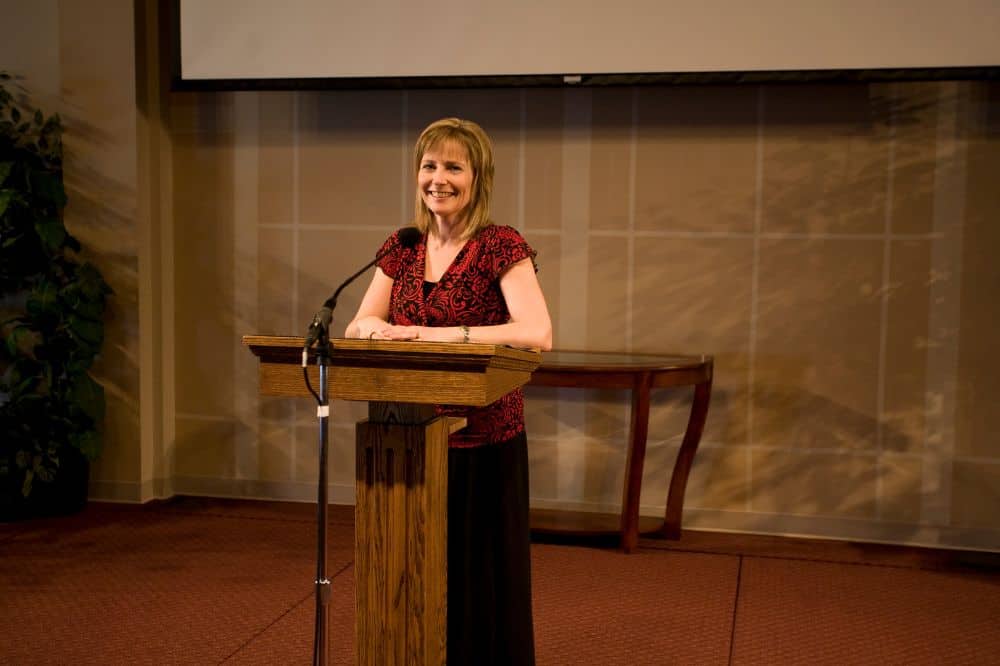 Elegant woman speaking at a church or religious conference, standing behind a wooden podium with a microphone, representing AEGA Ministries International, Inc.